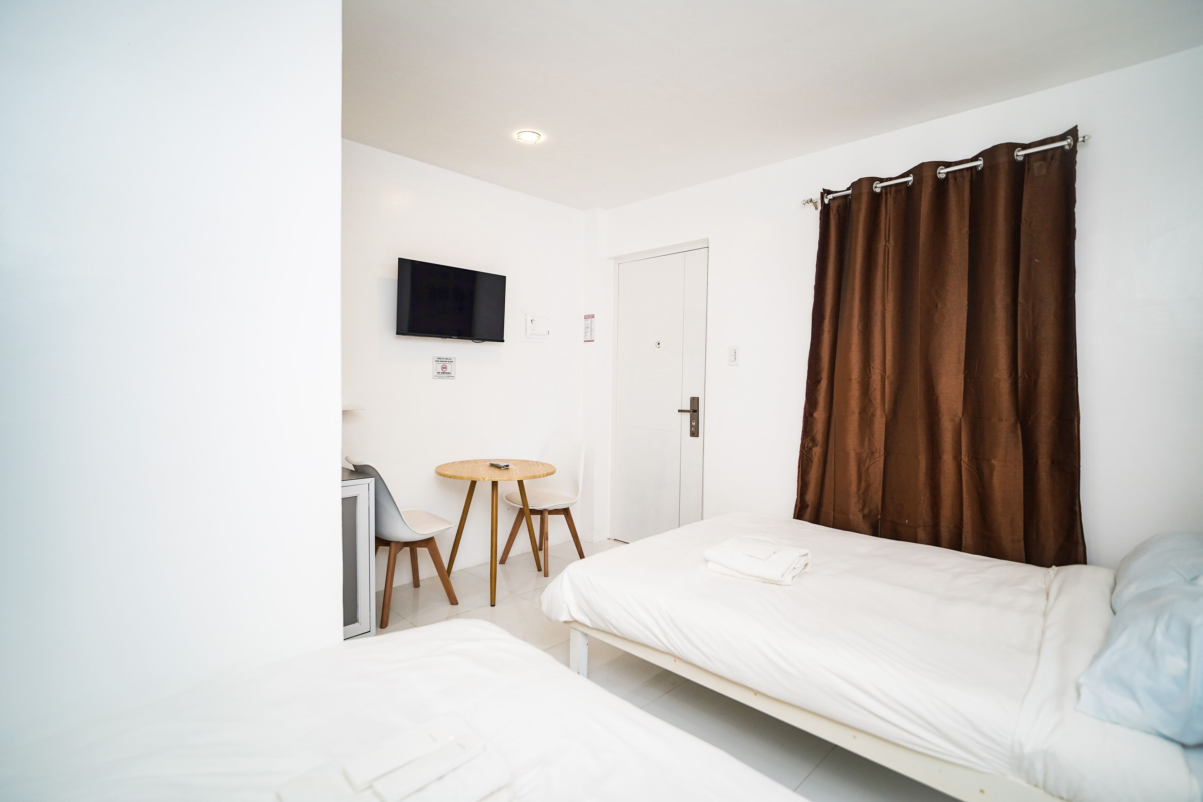 A hotel room with two beds, a TV mounted on the wall, a small round table with chairs, and a door with a brown curtain.