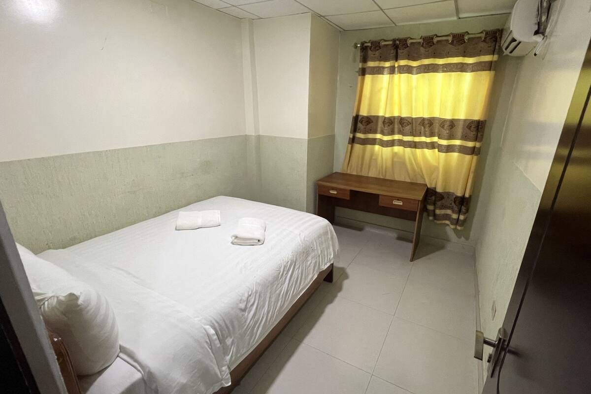 A small, clean room with a single bed, a wooden desk, and a window with curtains.