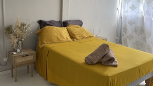 Ein Schlafzimmer mit einem Bett, einem Nachttisch, einer Vase mit Trockenblumen und einem Fenster mit Vorhängen.