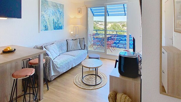 A modern, open-plan living space with a kitchen area, a dining table with stools, a sofa, and a large window offering a view of the outdoors. - 01426 _ Apartment 4 people, residence with pool, Lacanau Ocean (LACANAU OCEAN)