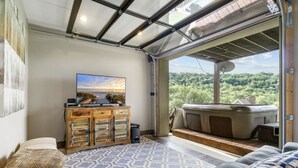 A modern living room with a large TV, a wooden cabinet, and a cozy seating area with a view of a forested landscape. - Tranquil Waters Retreat: Hot Tub, Pool & Lake Views (Smithville)