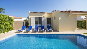 Une piscine entourée de chaises longues bleues, un espace extérieur aménagé et couvert, et un bâtiment doté d’un toit en tuiles.