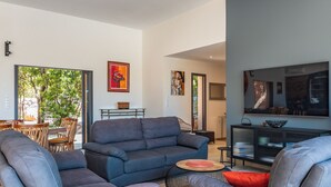 A modern living room with a grey sofa, a coffee table, and a TV. There is a dining area with a table and chairs visible through an open door.