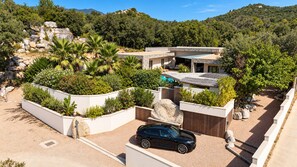 A modern house with a car parked in the driveway, surrounded by landscaped gardens and a hilly backdrop.