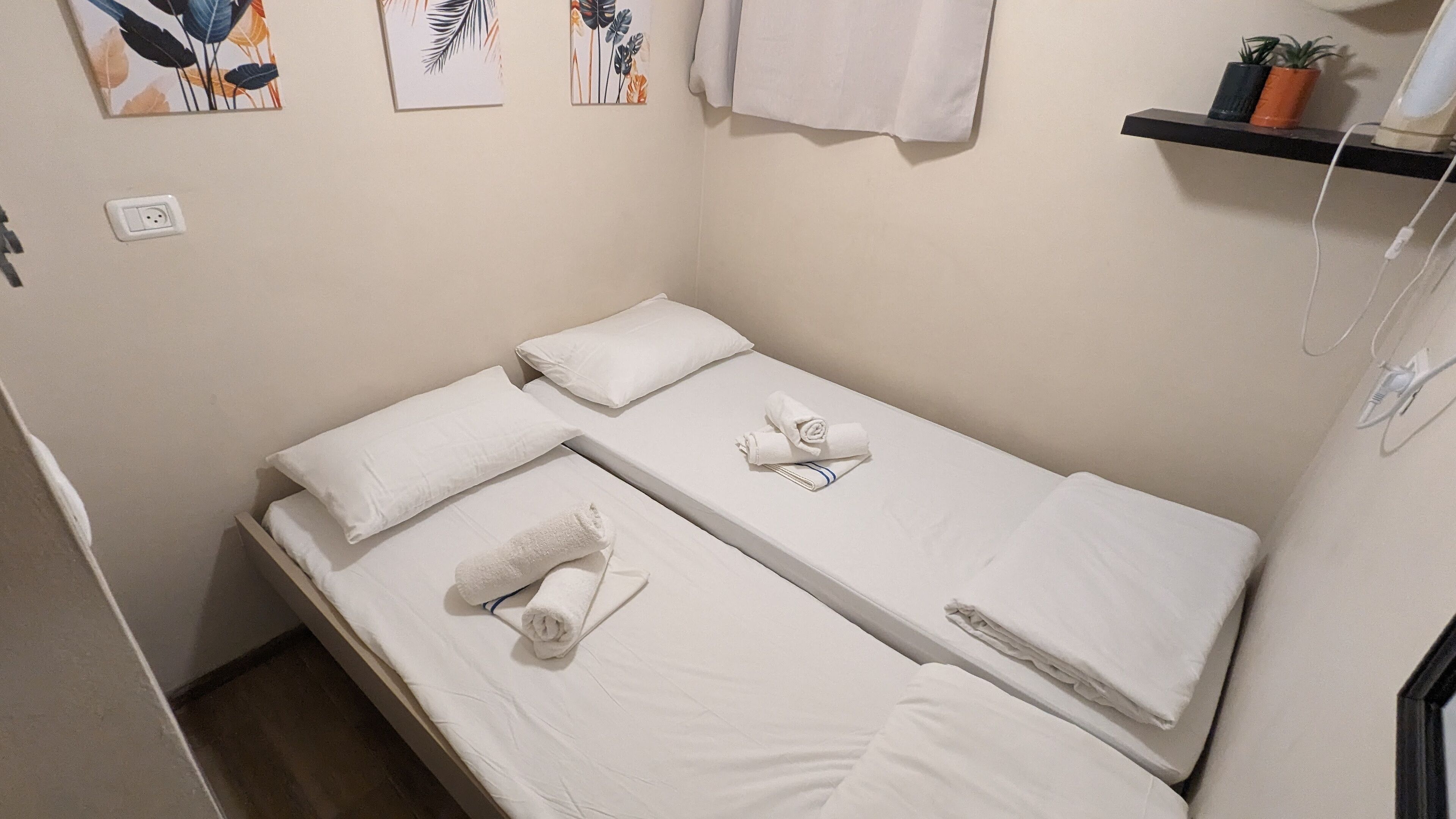 A small, neatly made bed with white linens and towels, a power outlet, and a shelf with two potted plants.