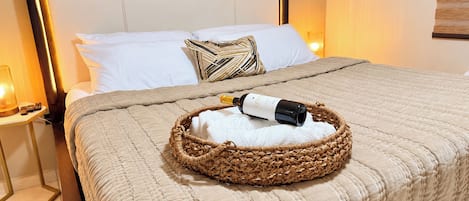 Een net opgemaakt bed met een fles wijn, een geweven mand en een witte handdoek.