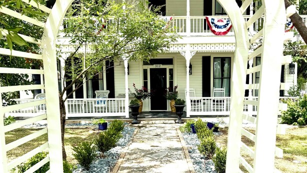 A white wooden porch with a number 312 sign, surrounded by a white picket fence, leading to a house with a black door.
