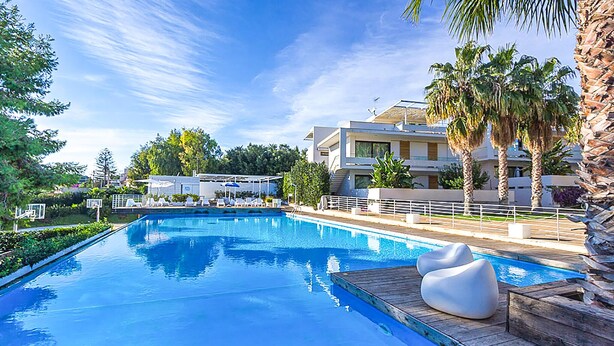 Une piscine dotée d’une terrasse en bois et de chaises longues, entourée de palmiers et bordée d’un bâtiment moderne avec des balcons.