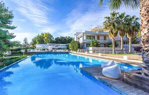 Una piscina con cubierta de madera y sillas de descanso, rodeada de palmeras y un edificio moderno con balcones.
