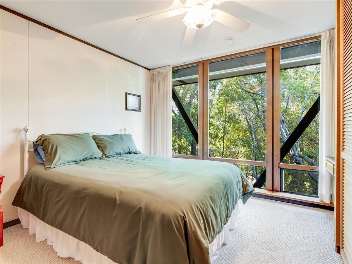 A bedroom with a bed, a ceiling fan, a window with a view of trees, and a red chair.