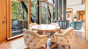 A modern dining area with a round table and four chairs, large windows offering a view of trees, and a fireplace in the background.