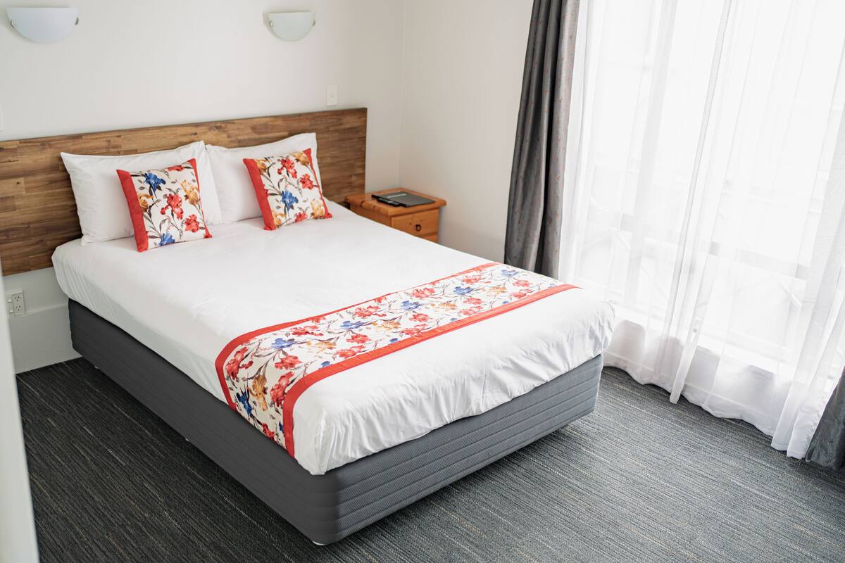 Une chambre d’hôtel avec un grand lit, une tête de lit en bois, une table de chevet et une fenêtre ornée de rideaux légers.