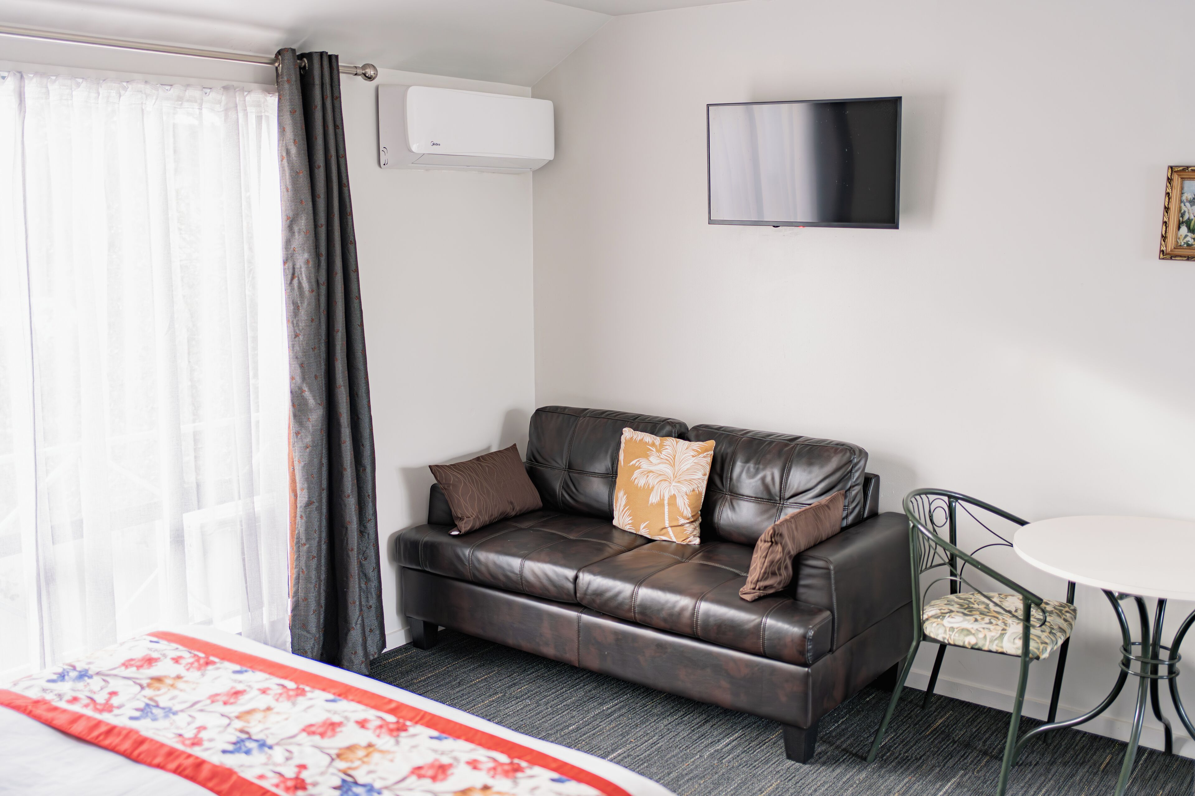 Une chambre d’hôtel moderne avec un canapé en cuir, une petite table ronde, un téléviseur à écran plat et un lit recouvert d’une courtepointe à motifs floraux.