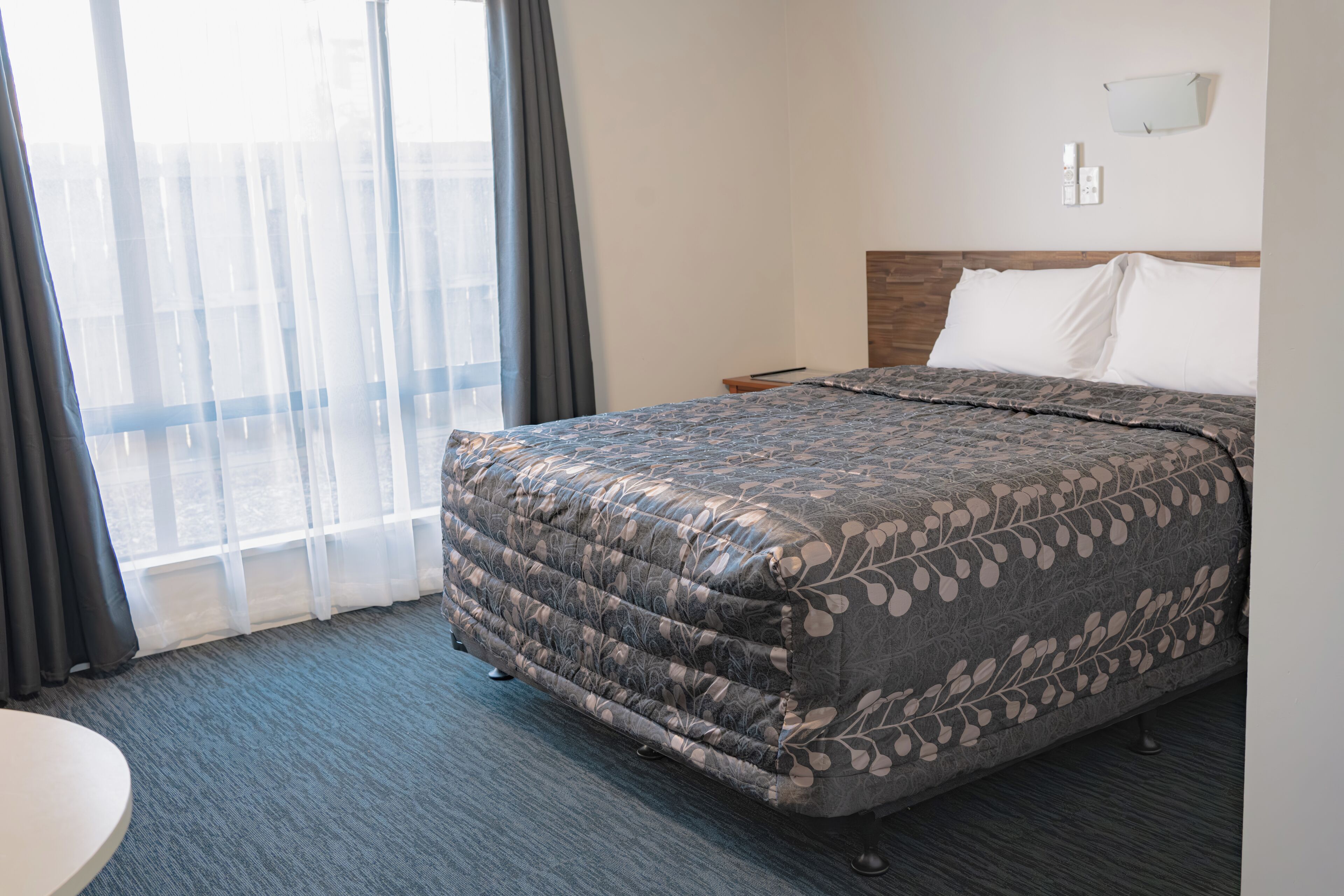A hotel room with a bed, a round table, a window with curtains, and a wall-mounted light fixture.