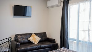 A room with a black leather sofa, a television mounted on the wall, and a window with sheer curtains. - Samhil Motor Lodge (Christchurch)