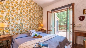 A bedroom with a large bed, a nightstand, a bedside table, a window with a view of greenery, and a door leading to an outdoor area.