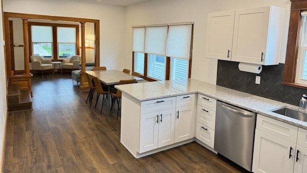 A modern kitchen with white cabinets, a granite countertop, and a dishwasher. There is a dining area with a table and chairs, and a view of the outdoors through large windows.