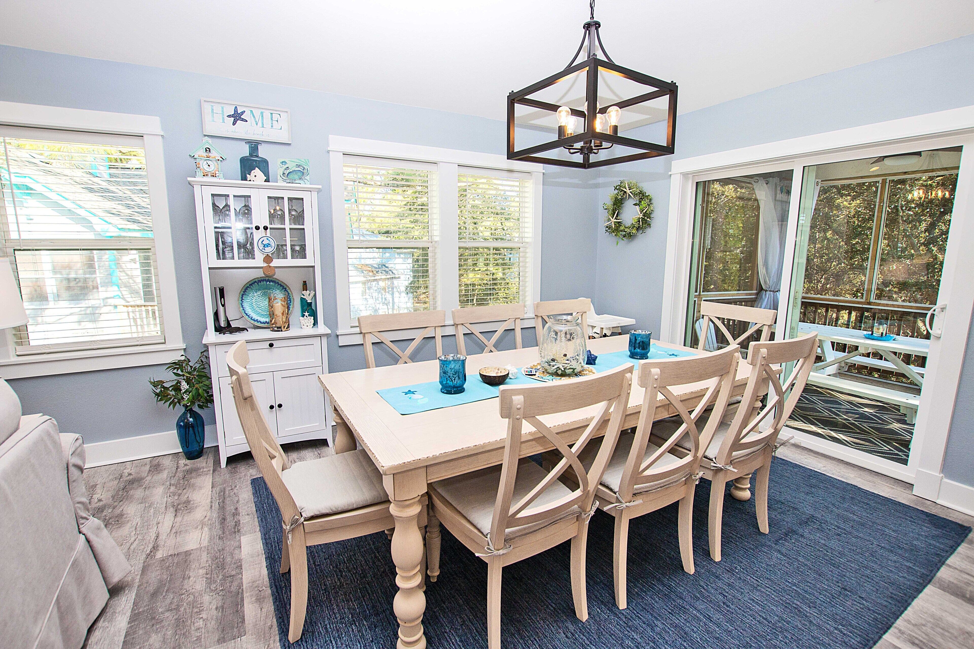 A dining area with a table set for six, surrounded by wooden chairs, a chandelier, and a view of the outdoors through large windows.