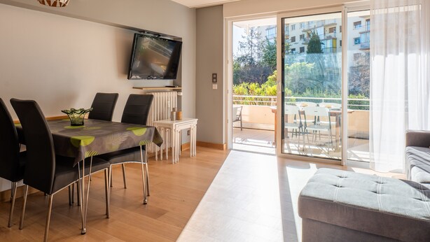A modern living room with a dining area, a flat-screen TV mounted on the wall, and a sliding glass door leading to an outdoor balcony.