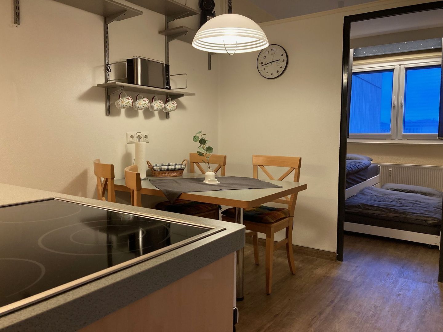 A compact kitchen with a stove, microwave, and hanging mugs. There's a dining table with chairs and a clock on the wall. A window reveals a cityscape at dusk.