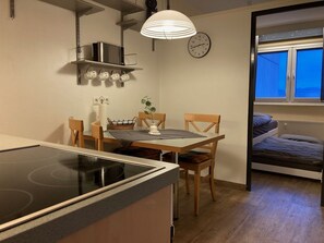 A compact kitchen with a stove, microwave, and hanging mugs. There's a dining table with chairs and a clock on the wall. A window reveals a cityscape at dusk.
