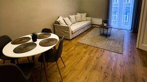 A modern living room with a white sofa, a dining table with placemats, a ceiling fan, and a view of the city through large windows. - CITY CENTER 2! A Cozy and Modern 2BR in the city center near New York Cafe! (Budapest)