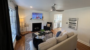 Una sala de estar con chimenea, televisor, ventilador de techo y vista a la cocina.