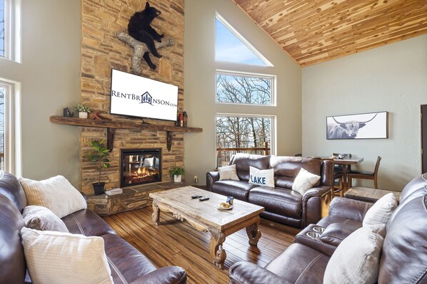 A spacious living room with a stone fireplace, leather sofas, a wooden coffee table, and a mounted TV. - Scenic Table Rock Shoreline w/ Access, Free Daily Tickets & Shared Amenities (Branson)