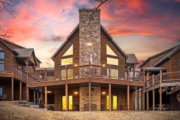 A two-story log cabin with a stone chimney, large windows, and a balcony with railings.
