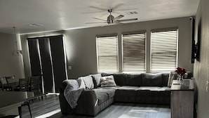 Un salon spacieux avec un canapé d’angle, un coin repas comprenant une table et des chaises, et un ventilateur de plafond.