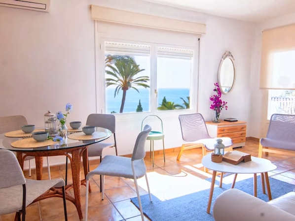 A modern living room with a dining area, a large window with a view of the sea, and a small side table with a mirror.
