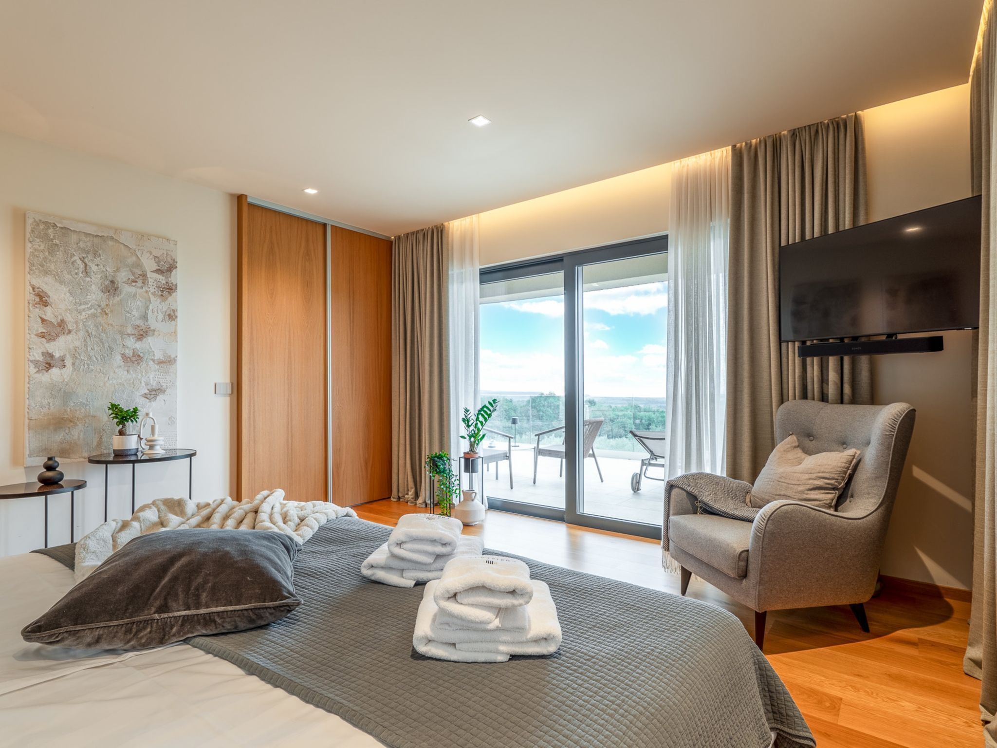 A modern hotel room with a large bed, a seating area with a chair, a flat-screen TV, and a balcony with a view.