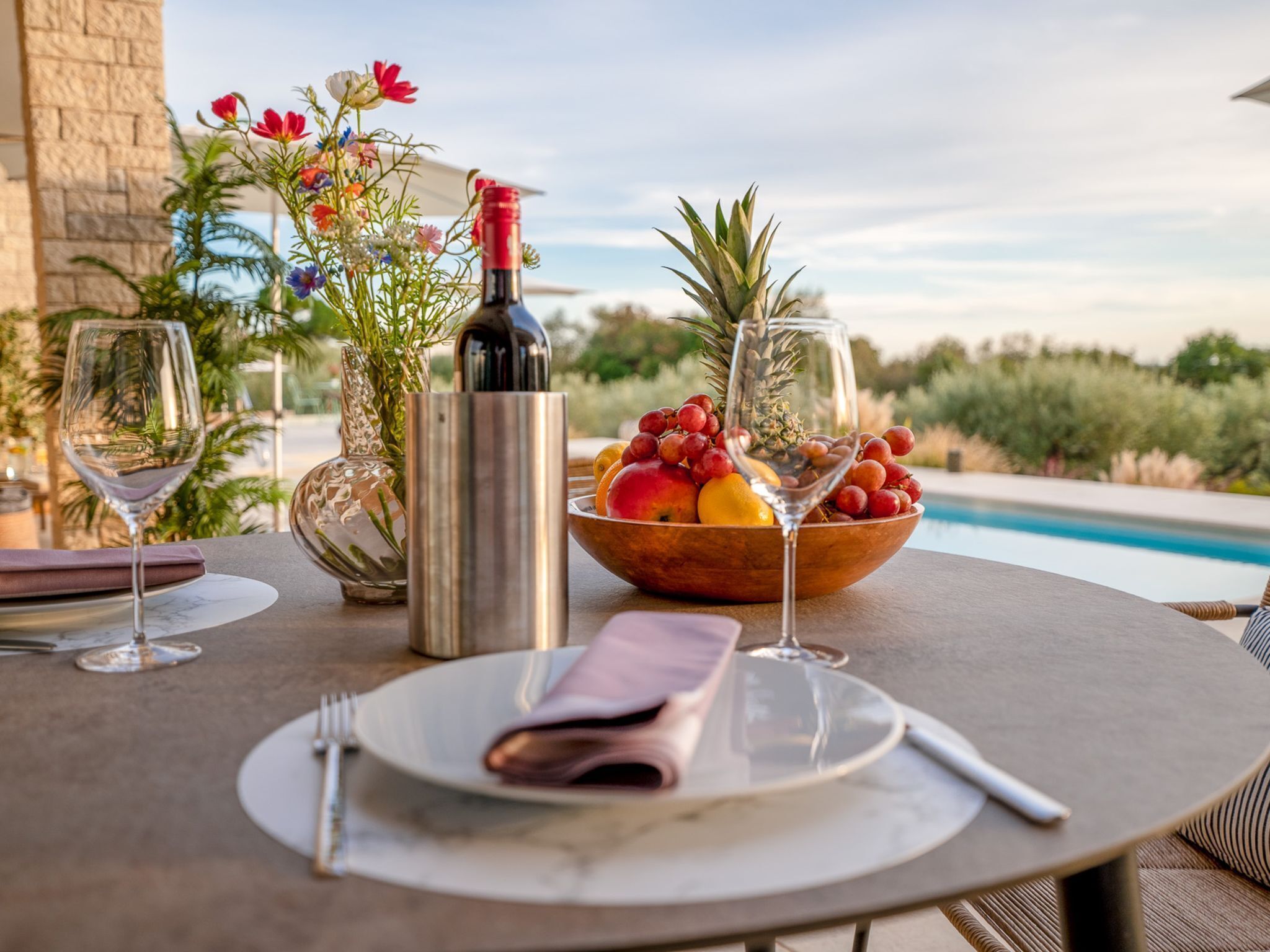 A table set with a bottle, a bowl of fruit, and two wine glasses, overlooking a pool.