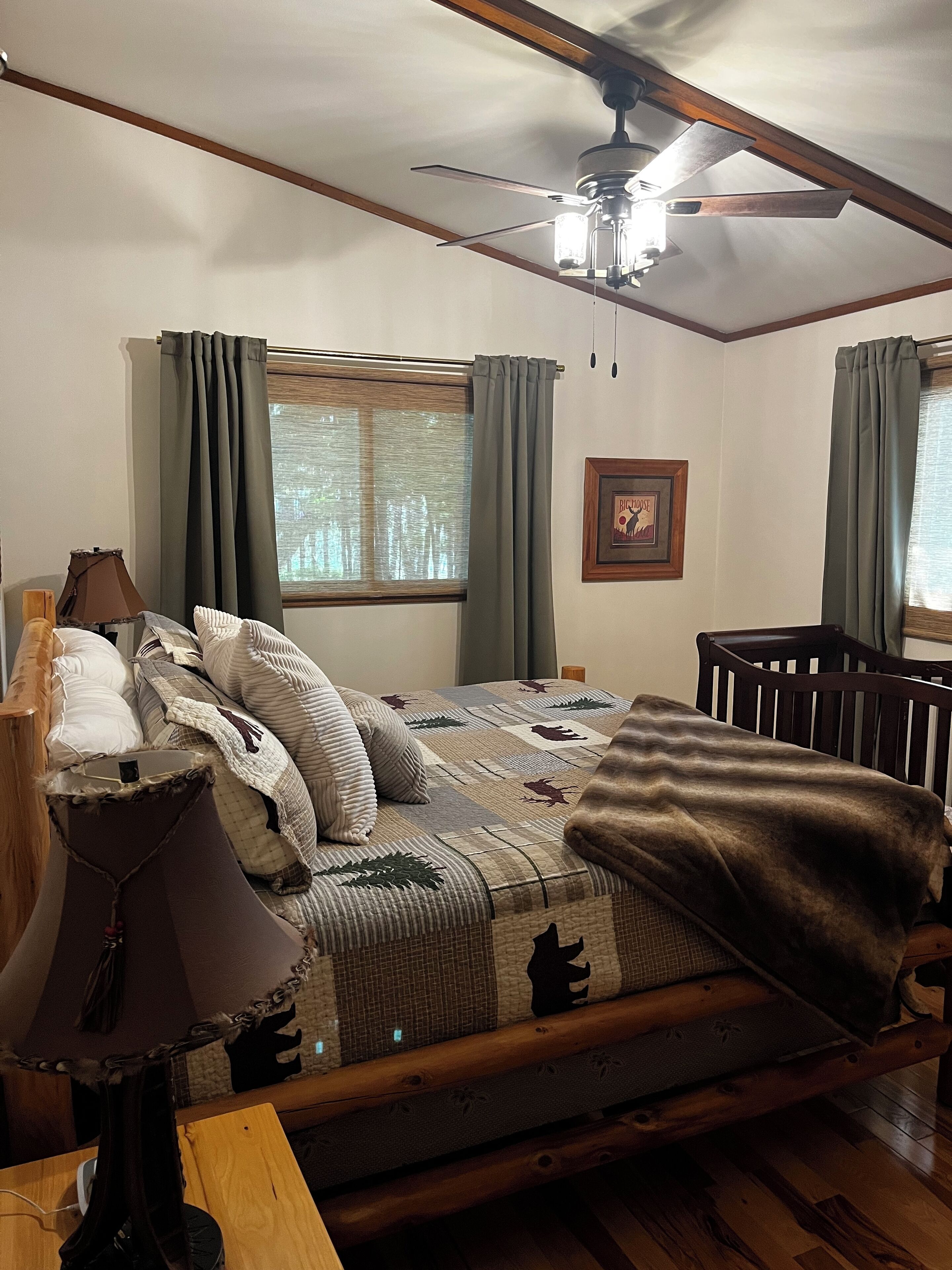 A bedroom with a wooden bed, a ceiling fan, a window with curtains, a framed picture on the wall, and a lamp on a bedside table.