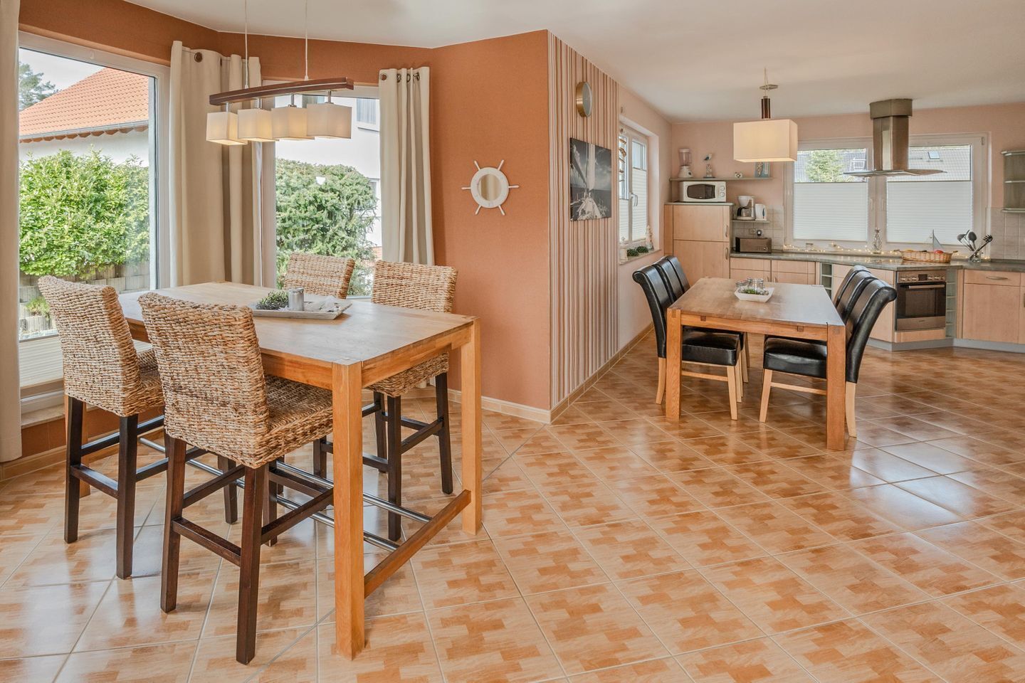 A dining area with a wooden table and wicker chairs, a kitchen with modern appliances, and a view of greenery outside.