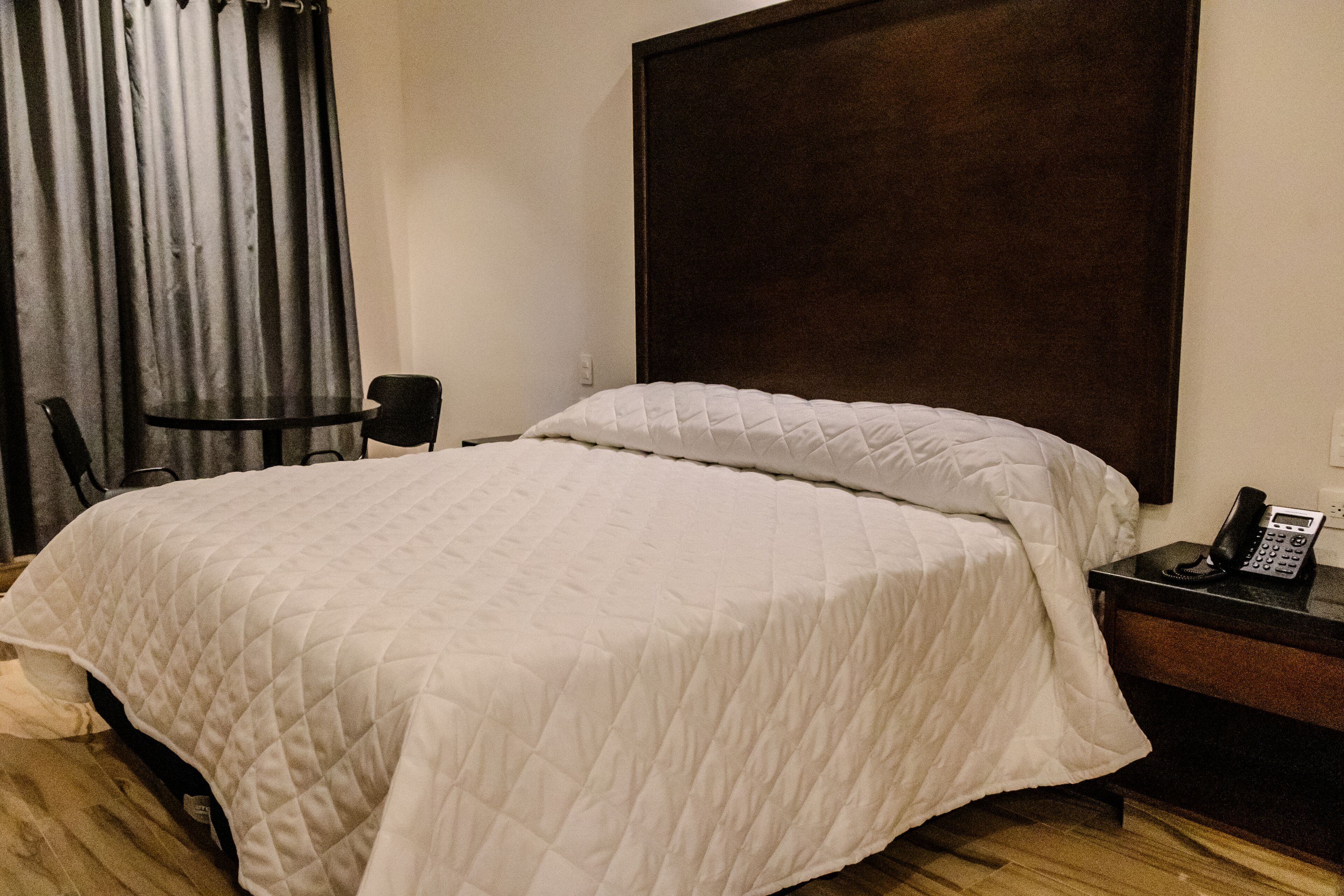 A neatly made bed with a white quilt, a wooden headboard, a bedside table with a phone, and a chair.