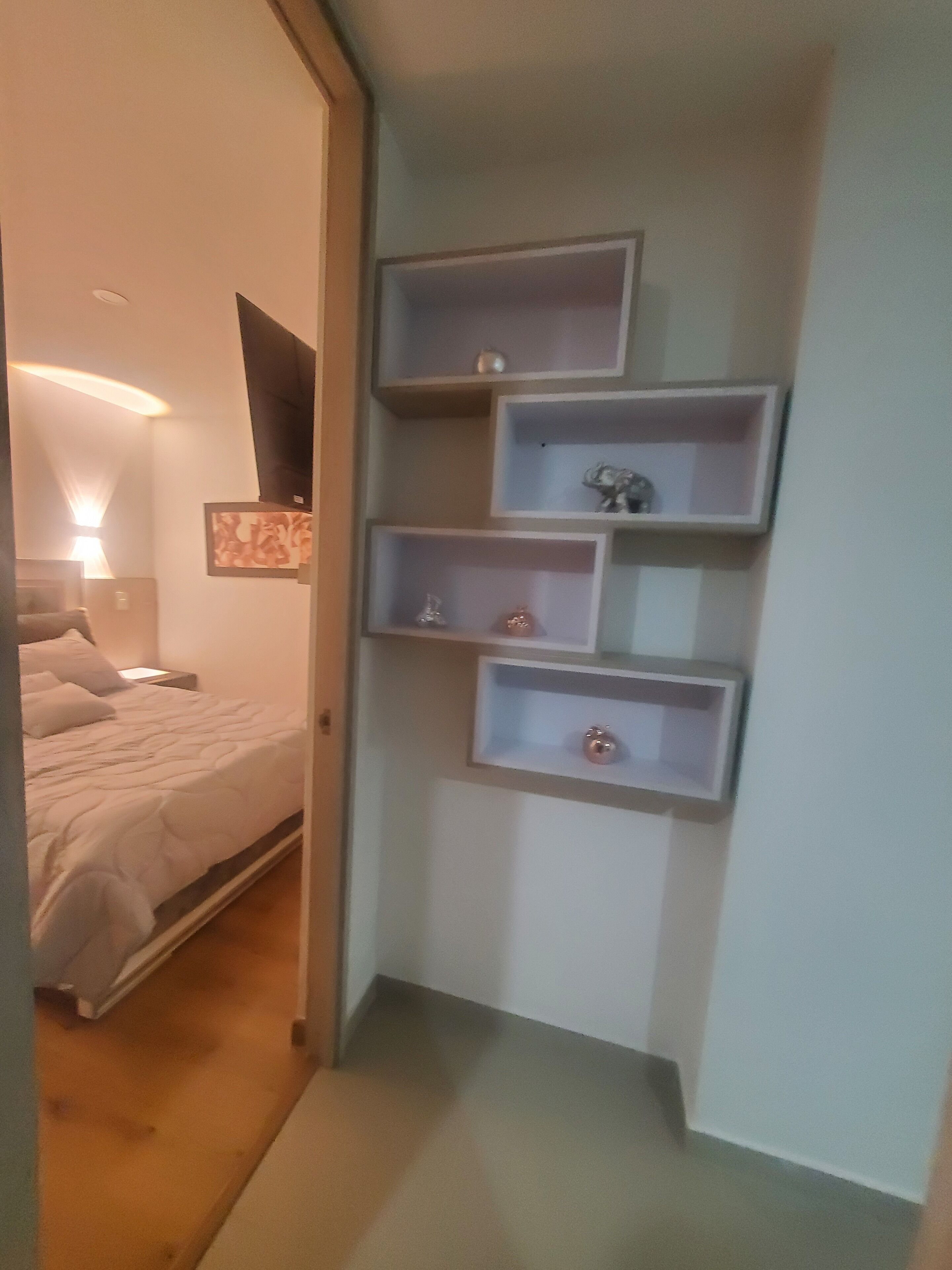 A bedroom with a bed, bedside table, lamp, and a wall-mounted shelf unit with decorative items.