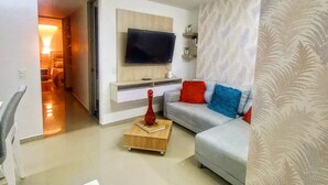 Una sala de estar moderna con un televisor de pantalla plana montado en un panel de madera, un sofá gris con cojines de colores, una mesita de centro de madera y una cortina estampada.