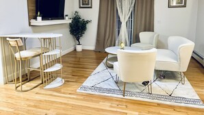 A modern living room with a white sofa, a round coffee table, and a dining area with a white table and gold chairs. There is a large window with curtains, a potted plant, and framed artwork on the walls. - Beautiful two-story home close to downtown worcester 5beds-4bedroom (Worcester)
