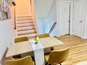 Un comedor con una mesa de madera y cuatro sillas amarillas, un globo como adorno central, una escalera con escalones de madera y una puerta blanca con un número.