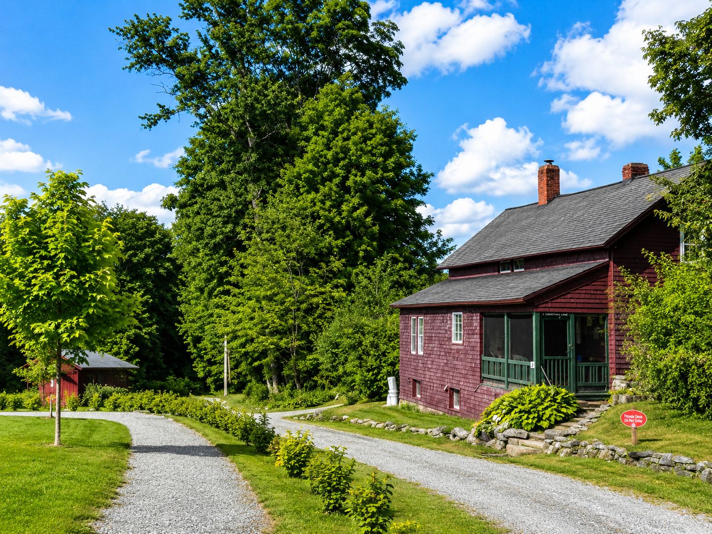 Une maison en bois rouge, avec une allée de gravier menant à une porte verte, entourée d’une végétation luxuriante et d’arbres.