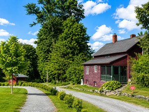 A red wooden house with a gravel driveway leading to a green door, surrounded by lush greenery and trees.