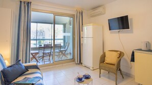 Una habitación con sofá, una silla de mimbre, una mesita con tazas, un refrigerador, un televisor montado en la pared y una puerta corrediza de vidrio que da a un área exterior.