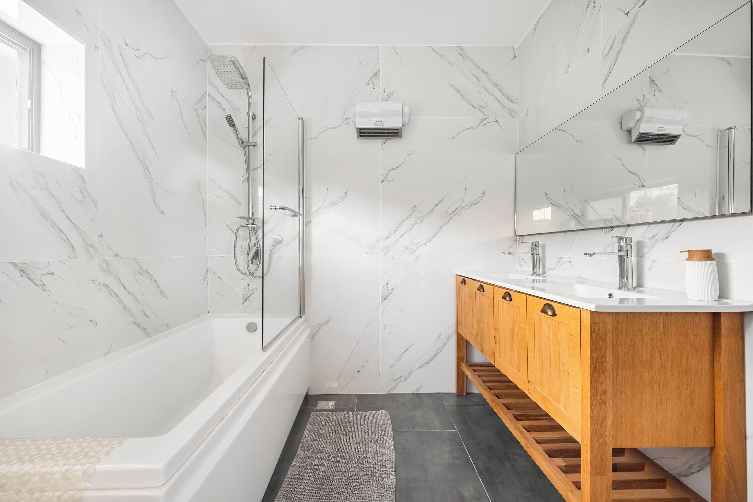A modern bathroom with a marble wall, a wooden vanity, a large mirror, and a shower area.