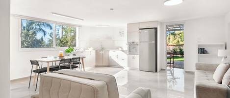 A modern, well-lit living area with a sofa, a dining table with chairs, a kitchen with a refrigerator, and large windows offering a view of the outdoors.