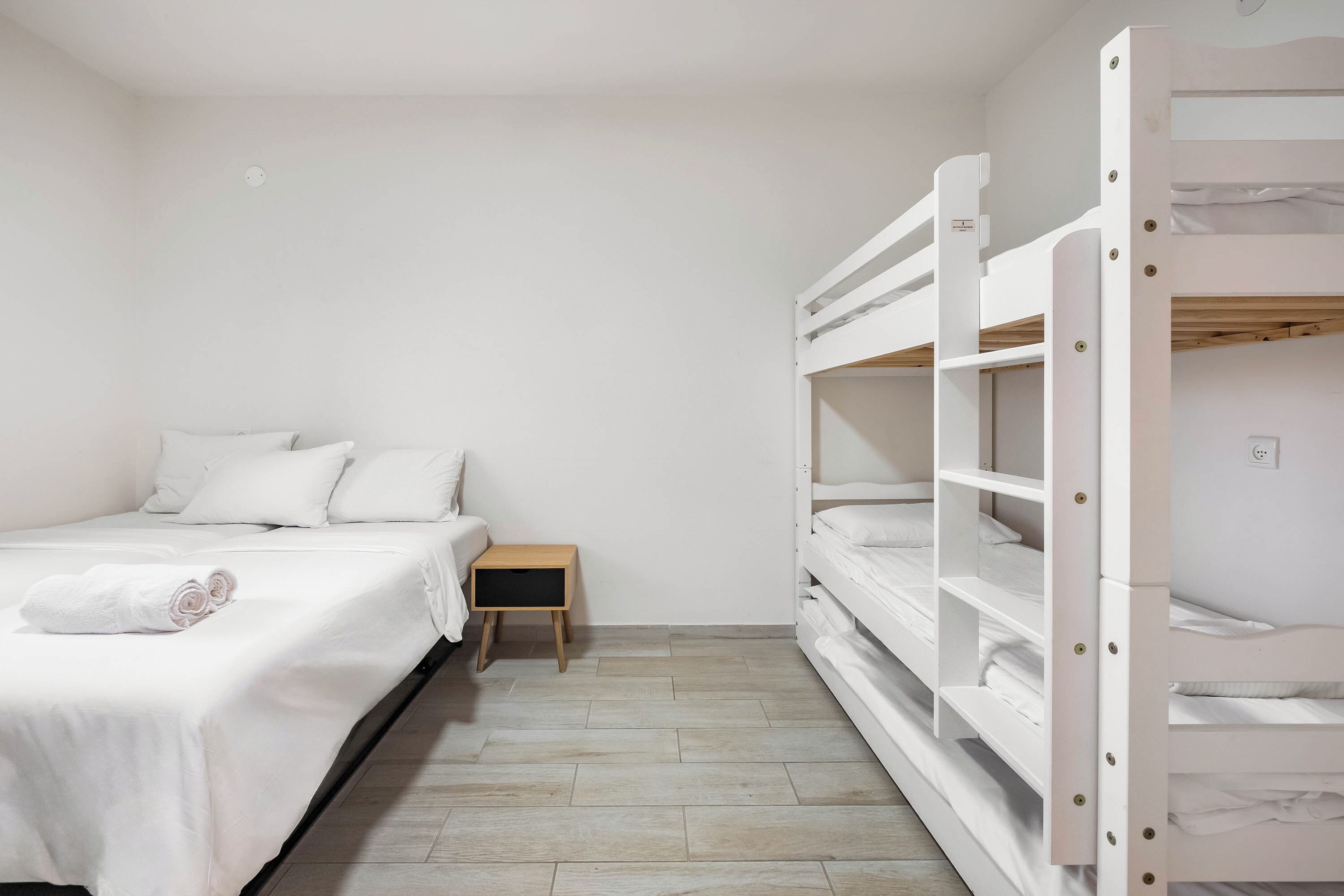 Un lit simple avec une table de chevet en bois, un mur blanc et un lit superposé avec une literie blanche.