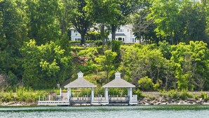 A gazebo on a dock with a view of a white house and lush greenery in the background.