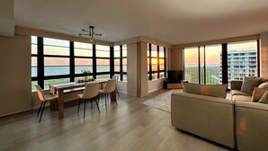 A spacious living room with large windows offering ocean views, a dining area with a table and chairs, and a comfortable sofa.