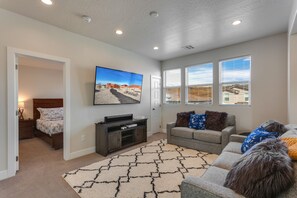 A living room with a flat-screen TV, a grey sofa, a patterned rug, and a window with blinds.