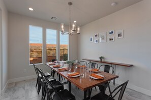 A dining area with a long table set for four, chairs, a chandelier, and framed pictures on the wall.